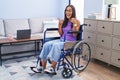 Young hispanic woman sitting on wheelchair at home success sign doing positive gesture with hand, thumbs up smiling and happy Royalty Free Stock Photo