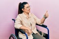 Young hispanic woman sitting on wheelchair looking proud, smiling doing thumbs up gesture to the side