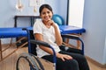 Young hispanic woman sitting on wheelchair at physiotherapy clinic smiling looking confident at the camera with crossed arms and Royalty Free Stock Photo
