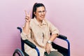 Young hispanic woman sitting on wheelchair smiling looking to the camera showing fingers doing victory sign Royalty Free Stock Photo