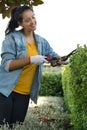 Young hispanic woman smiling and working on the yard cutting bush with hedge shear