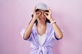 Young hispanic woman standing over pink background wearing hat doing ok gesture like binoculars sticking tongue out, eyes looking Royalty Free Stock Photo