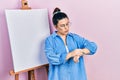 Young hispanic woman standing by painter easel stand checking the time on wrist watch, relaxed and confident Royalty Free Stock Photo