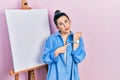 Young hispanic woman standing by painter easel stand in hurry pointing to watch time, impatience, looking at the camera with Royalty Free Stock Photo
