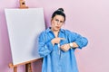 Young hispanic woman standing by painter easel stand in hurry pointing to watch time, impatience, upset and angry for deadline Royalty Free Stock Photo