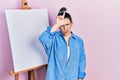 Young hispanic woman standing by painter easel stand making fun of people with fingers on forehead doing loser gesture mocking and Royalty Free Stock Photo