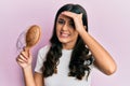 Young hispanic woman styling hair using comb stressed and frustrated with hand on head, surprised and angry face Royalty Free Stock Photo