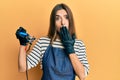 Young hispanic woman tattoo artist wearing professional uniform and gloves covering mouth with hand, shocked and afraid for