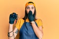 Young hispanic woman tattoo artist wearing professional uniform and gloves holding tattooer machine covering mouth with hand,