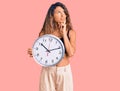 Young hispanic woman with tattoo holding big clock serious face thinking about question with hand on chin, thoughtful about Royalty Free Stock Photo