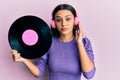 Young hispanic woman using headphones holding vinyl disc relaxed with serious expression on face