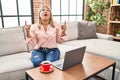 Young hispanic woman using laptop sitting on the sofa at home amazed and surprised looking up and pointing with fingers and raised Royalty Free Stock Photo