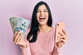 Young hispanic woman using smartphone holding united states dollar banknotes smiling and laughing hard out loud because funny