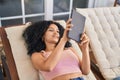 Young hispanic woman using touchpad lying on deck chair at home terrace