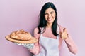 Young hispanic woman wearing baker uniform holding homemade bread smiling happy and positive, thumb up doing excellent and Royalty Free Stock Photo