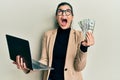 Young hispanic woman wearing business style holding laptop and 50 euros angry and mad screaming frustrated and furious, shouting Royalty Free Stock Photo