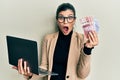 Young hispanic woman wearing business style holding laptop and swedish krone afraid and shocked with surprise and amazed