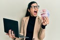 Young hispanic woman wearing business style holding laptop and swedish krone angry and mad screaming frustrated and furious,