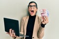 Young hispanic woman wearing business style holding laptop and swedish krone angry and mad screaming frustrated and furious,