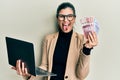 Young hispanic woman wearing business style holding laptop and swedish krone sticking tongue out happy with funny expression