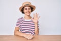 Young hispanic woman wearing casual clothes and hat sitting on the table showing and pointing up with fingers number five while Royalty Free Stock Photo