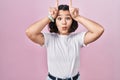 Young hispanic woman wearing casual white t shirt over pink background doing funny gesture with finger over head as bull horns Royalty Free Stock Photo