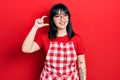 Young hispanic woman wearing cook apron and glasses smiling and confident gesturing with hand doing small size sign with fingers Royalty Free Stock Photo