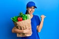 Young hispanic woman wearing courier uniform with groceries from supermarket pointing thumb up to the side smiling happy with open Royalty Free Stock Photo