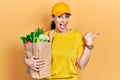 Young hispanic woman wearing courier uniform with groceries from supermarket pointing thumb up to the side smiling happy with open Royalty Free Stock Photo