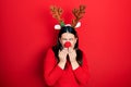 Young hispanic woman wearing deer christmas hat and red nose laughing and embarrassed giggle covering mouth with hands, gossip and
