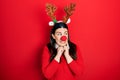 Young hispanic woman wearing deer christmas hat and red nose laughing nervous and excited with hands on chin looking to the side