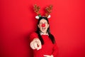 Young hispanic woman wearing deer christmas hat and red nose laughing at you, pointing finger to the camera with hand over body,