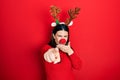 Young hispanic woman wearing deer christmas hat and red nose laughing at you, pointing finger to the camera with hand over mouth,