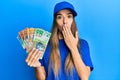 Young hispanic woman wearing delivery uniform and cap holding australian dollars covering mouth with hand, shocked and afraid for Royalty Free Stock Photo