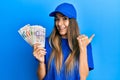 Young hispanic woman wearing delivery uniform and cap holding singapore dollars pointing thumb up to the side smiling happy with Royalty Free Stock Photo