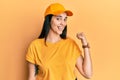 Young hispanic woman wearing delivery uniform and cap smiling with happy face looking and pointing to the side with thumb up Royalty Free Stock Photo