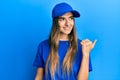Young hispanic woman wearing delivery uniform and cap smiling with happy face looking and pointing to the side with thumb up Royalty Free Stock Photo