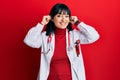 Young hispanic woman wearing doctor uniform and stethoscope smiling pulling ears with fingers, funny gesture