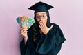 Young hispanic woman wearing graduation uniform holding australian dollars covering mouth with hand, shocked and afraid for Royalty Free Stock Photo