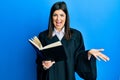 Young hispanic woman wearing judge uniform reading book celebrating achievement with happy smile and winner expression with raised