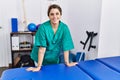 Young hispanic woman wearing physiotherapist uniform leaning on massage table at physiotherapy clinic Royalty Free Stock Photo