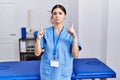 Young hispanic woman wearing physiotherapist uniform standing at clinic pointing up looking sad and upset, indicating direction