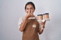 Young hispanic woman wearing professional waitress apron holding coffee smelling something stinky and disgusting, intolerable