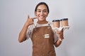 Young hispanic woman wearing professional waitress apron holding coffee smiling cheerful showing and pointing with fingers teeth