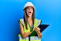 Young hispanic woman wearing safety helmet holding clipboard angry and mad screaming frustrated and furious, shouting with anger Royalty Free Stock Photo