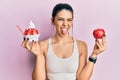 Young hispanic woman wearing sportswear holding apple and ice cream sticking tongue out happy with funny expression Royalty Free Stock Photo