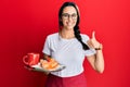 Young hispanic woman wearing waitress apron holding tray with breakfast smiling happy and positive, thumb up doing excellent and Royalty Free Stock Photo