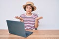 Young hispanic woman working with laptop sitting on the table clueless and confused expression with arms and hands raised Royalty Free Stock Photo