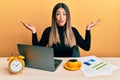 Young hispanic woman working at the office with laptop clueless and confused with open arms, no idea and doubtful face Royalty Free Stock Photo