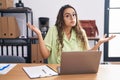 Young hispanic woman working at the office wearing glasses clueless and confused expression with arms and hands raised Royalty Free Stock Photo